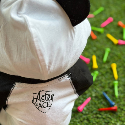 Close-up of the back of a baby hooded bodysuit with black bear ears and contrast stitching, featuring the Aster and Ace shield logo near the neckline, shown on grass with colorful golf tees.