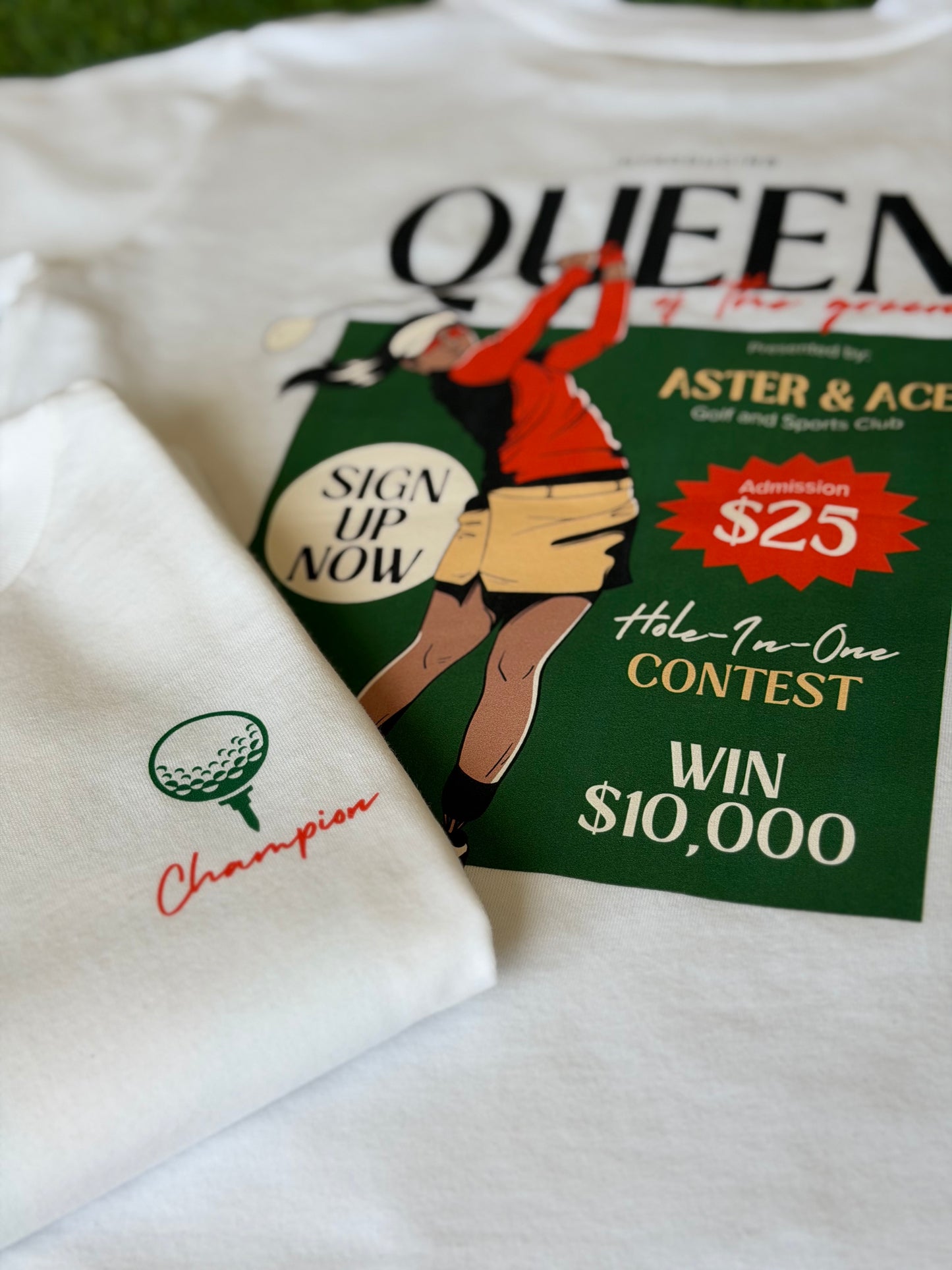 Two white golf themed shirts for girls. The first is folded to the side displaying the front left pocket design with a green golf ball and green tee above the word Champion written in orange script font. The second tshirt is to the side and behind the first and displays the large Queen of the green graphic on the back of the t-shirt.