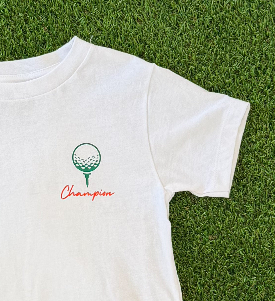 Front of boys’ white King of the Green golf t-shirt featuring a small green golf ball on a tee graphic with the word “Champion” in red script beneath it, shown on grass background.