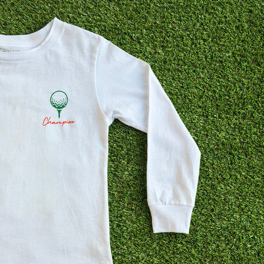 Front of long sleeve white golf t-shirt featuring a small green golf ball on a tee graphic with the word “Champion” in red script beneath it, displayed on a grass background.