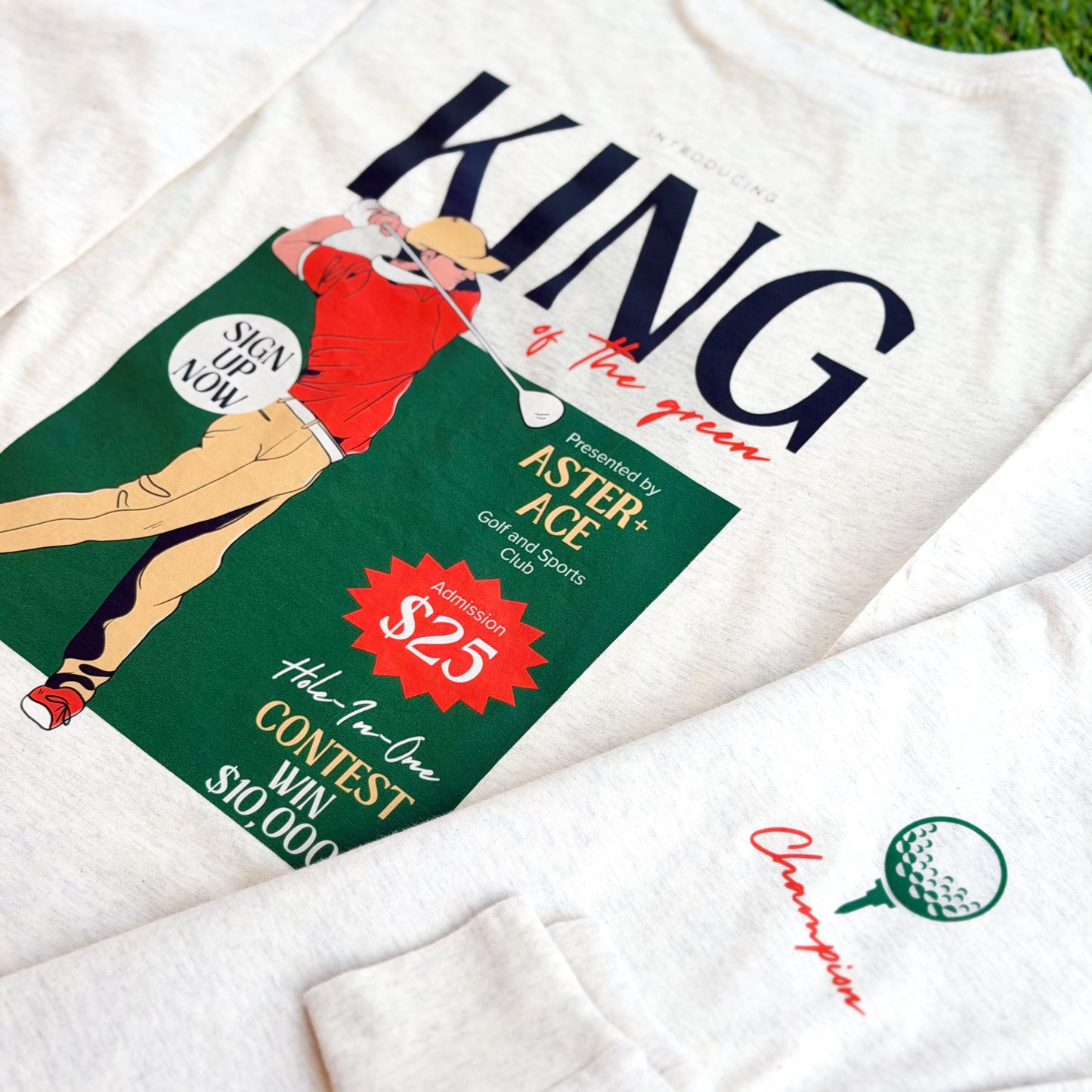 Front and back of a boys’ long sleeve heather beige golf t-shirt. The back features a colorful “King of the Green” graphic with a golfer in red and tan, and the front shows a small green golf ball on a tee graphic with the word “Champion” in orange script.