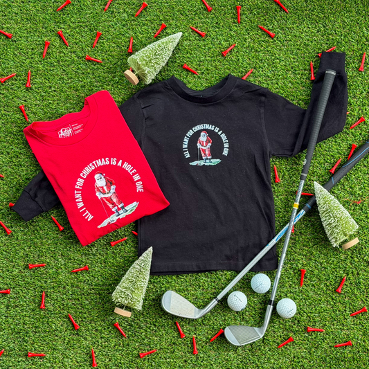 Two kids’ Christmas golf shirts, one red and one black, featuring a Santa golfing design with the text “All I Want for Christmas Is a Hole in One,” displayed on turf with golf clubs, tees, and small Christmas trees.