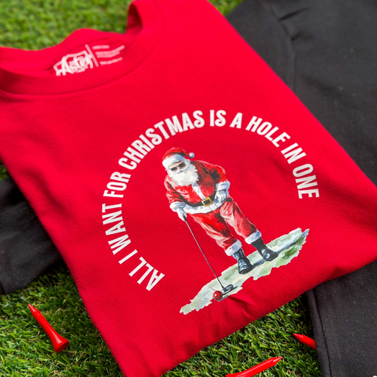 Close-up of a red kids’ Christmas golf t-shirt with a Santa golfing graphic and the words “All I Want for Christmas Is a Hole in One,” laid flat on green turf with red golf tees.