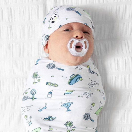 A newborn baby swaddled in a white muslin swaddle blanket with a colorful tennis-themed print, wearing a matching printed cap and having a white pacifier in their mouth, lying on a white textured blanket.
