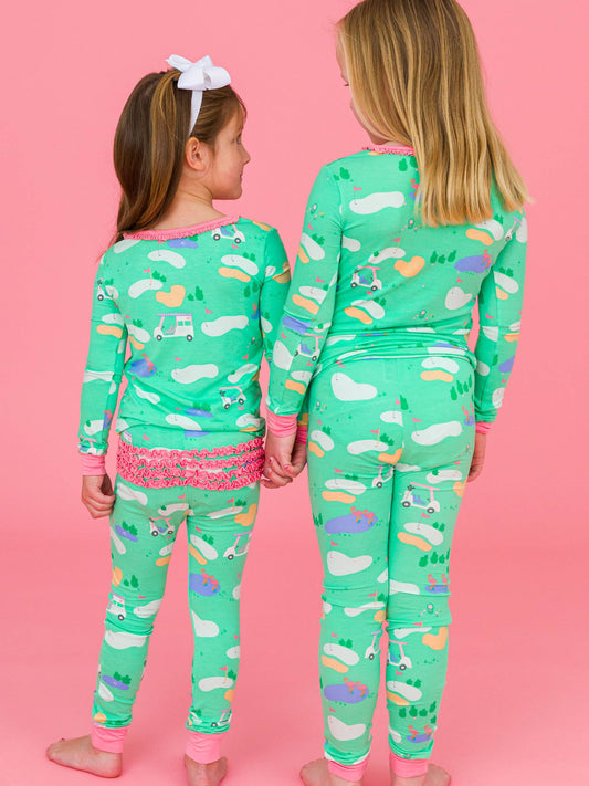 Toddler and girl wearing green golf course print pajamas. Back view. The toddler pjs have pink ruffles on the rear and the big girl pjs do not.