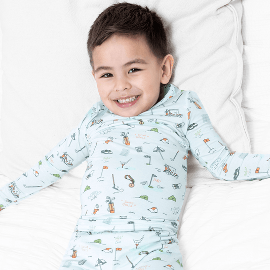 A smiling young boy lies on a white bed wearing a light blue long-sleeve pajama set with a colorful golf print. Text in the bottom right corner reads 'model: size worn 3T height 2' 10" weight 32 lbs'.