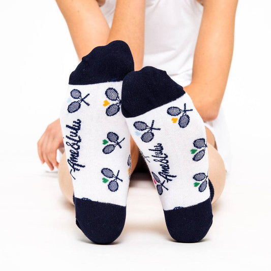 Person sitting with legs extended wearing white ankle socks with navy heel and toe, tennis racket pattern, and Ame & Lulu logo visible on the soles