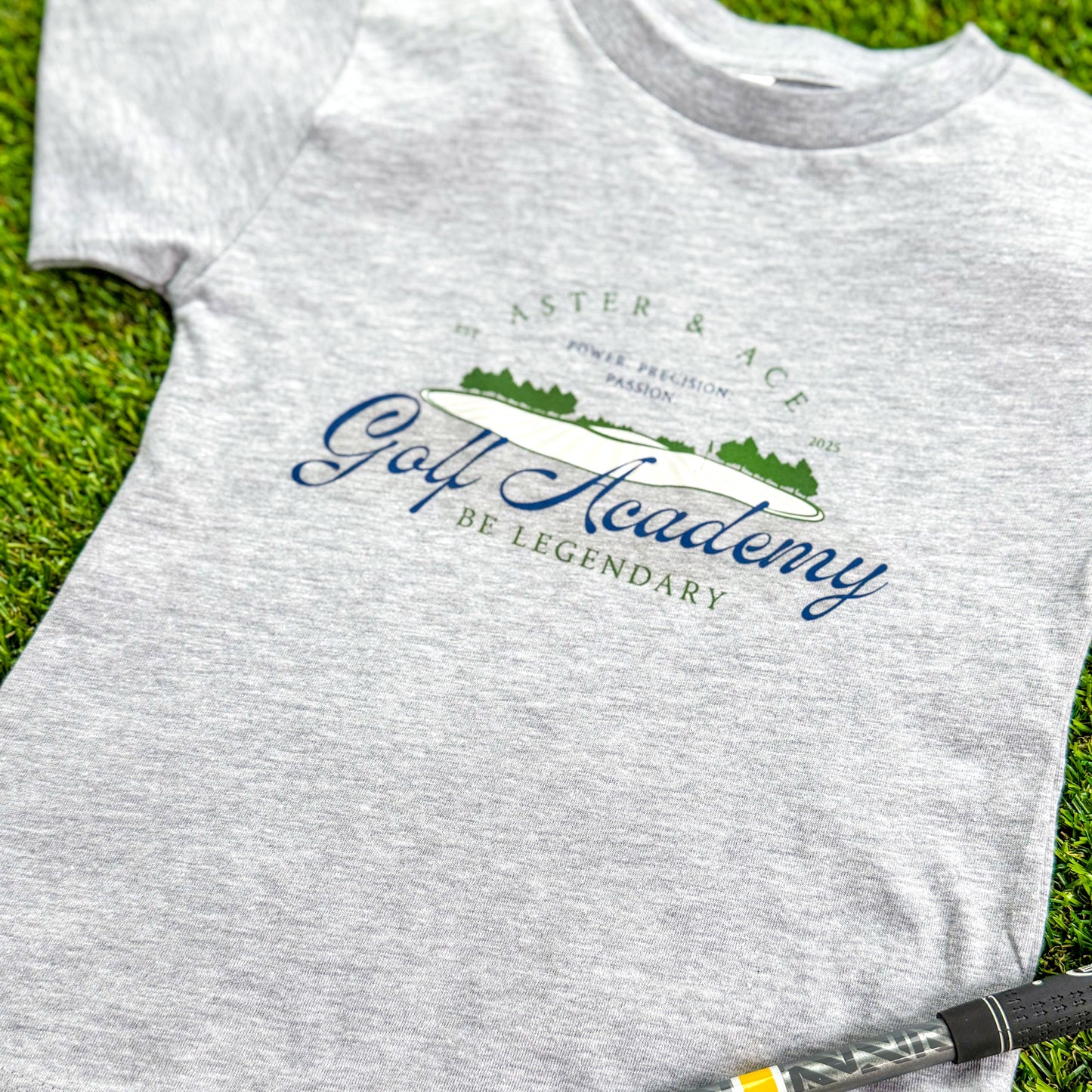 Aster and Ace Golf Academy kids graphic tee in heather grey styled on green turf with golf club detail.
