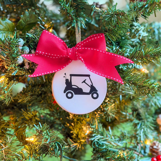 White ceramic ornament featuring a black golf cart silhouette, tied with a large red stitched ribbon and hanging on a decorated Christmas tree.