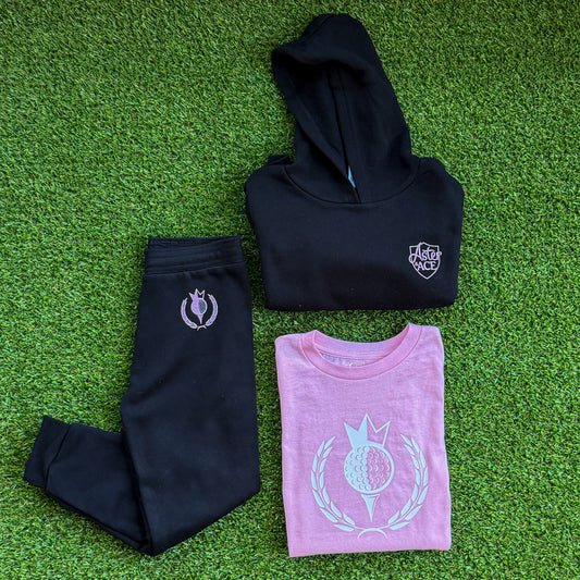 Black kids joggers and hoodie set by Aster and Ace with pink Royal-Tee golf crest, styled on turf with matching pink kids golf tee.
