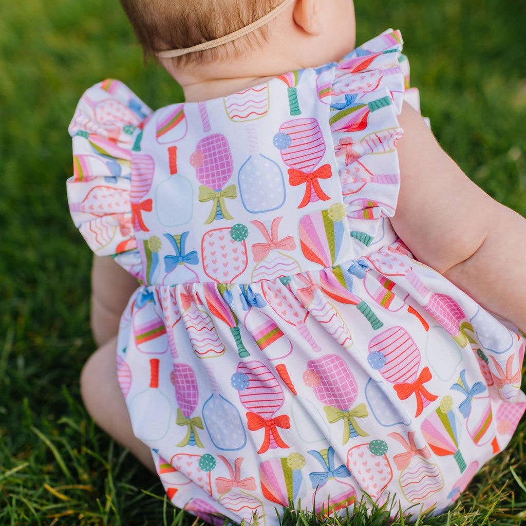 Baby sitting in grass wearing ruffle-sleeved romper with a colorful bow and pickleball paddle pattern.
