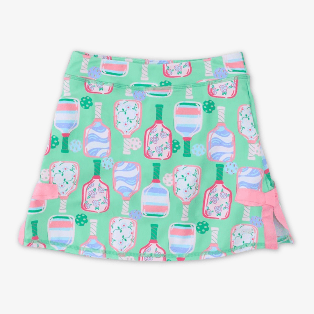 A girls' skort featuring a mint green background with a repeating pattern of pink and blue pickleball paddles, accented with pink side bows, shown on a plain light gray background.