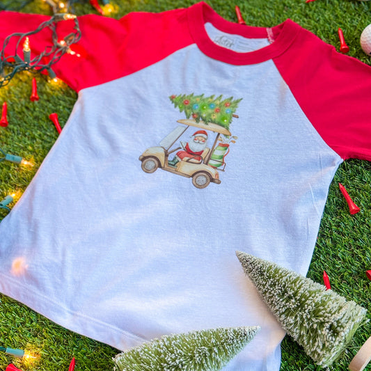 Kids’ red-and-white baseball tee featuring a festive golf cart graphic with Santa driving and a decorated Christmas tree on top, displayed on turf with holiday lights and mini bottlebrush trees.