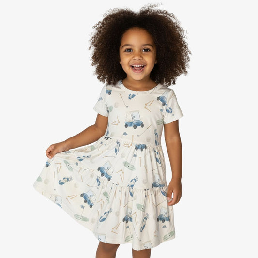 Smiling toddler wearing a short-sleeve golf-print dress with illustrated carts and clubs, photographed against a white background