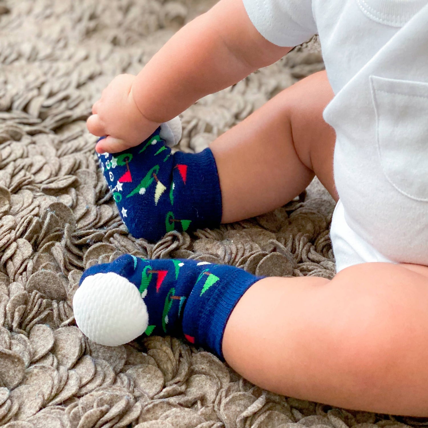 Baby sitting on a textured blanket wearing navy golf-themed rattle socks with white golf-ball toes.