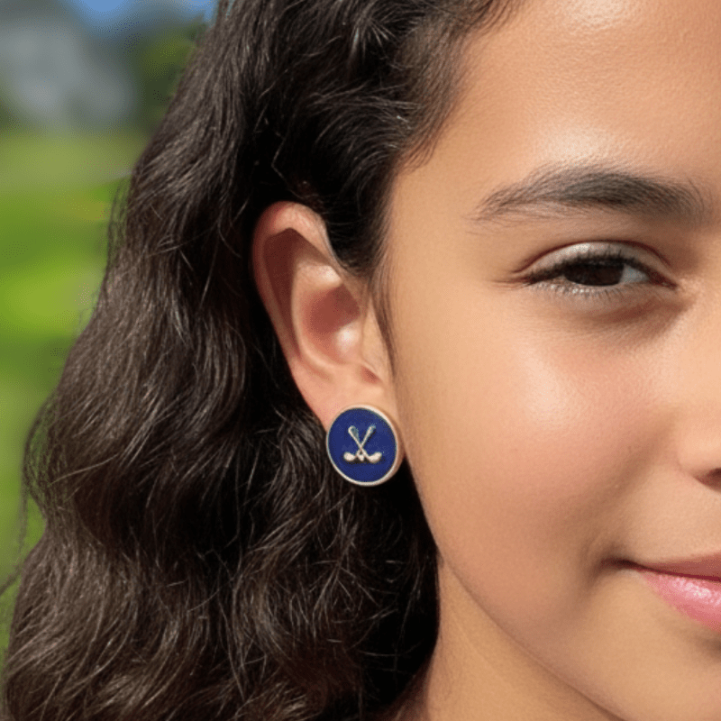Close-up of a young girl wearing Aster and Ace navy stud earrings with gold crossed golf clubs on a sunny golf course background.