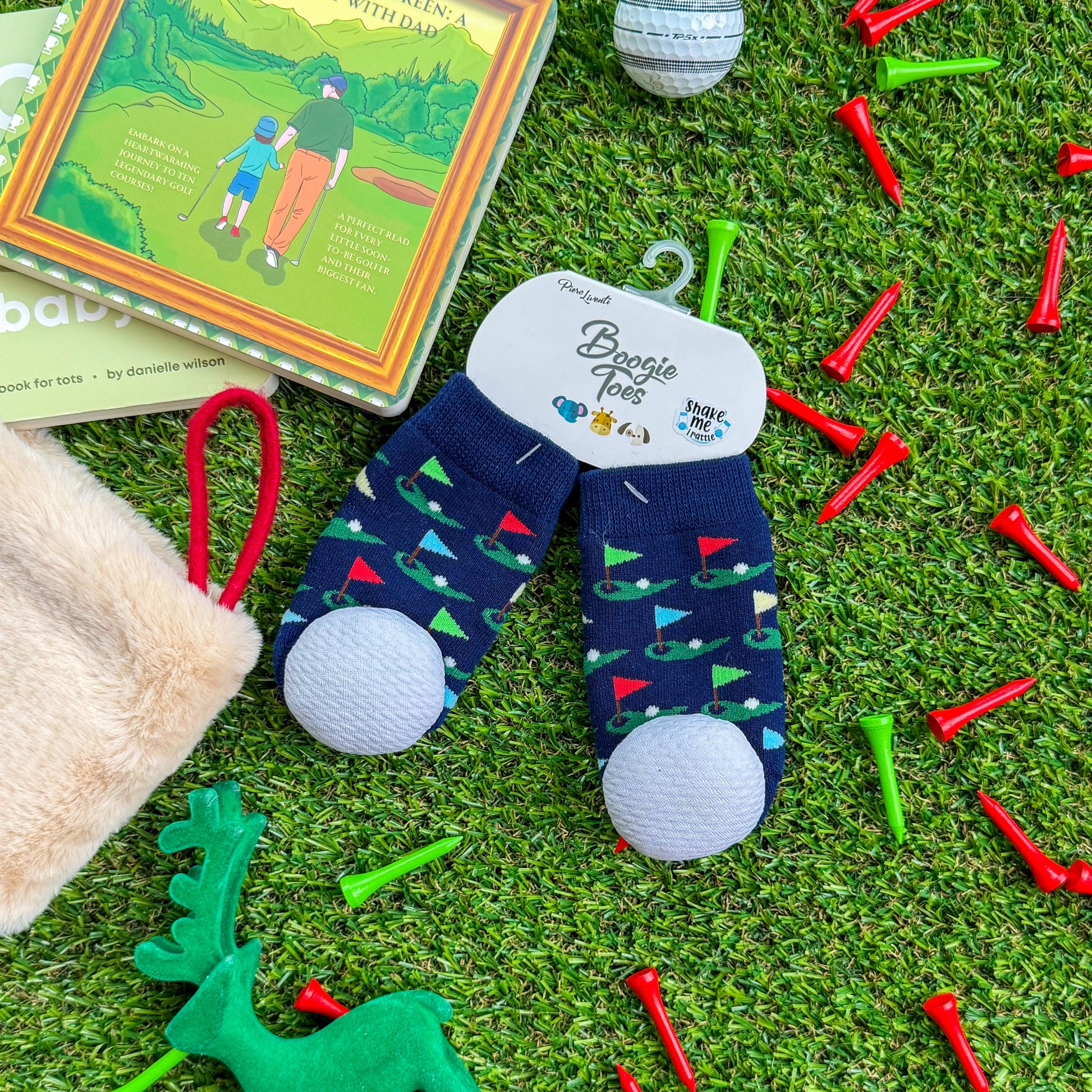 Boogie Toes baby rattle socks with golf flag and green print displayed on turf beside children's golf books, holiday stocking, and colorful golf tees—fun golf-themed stocking stuffer for babies and toddlers.