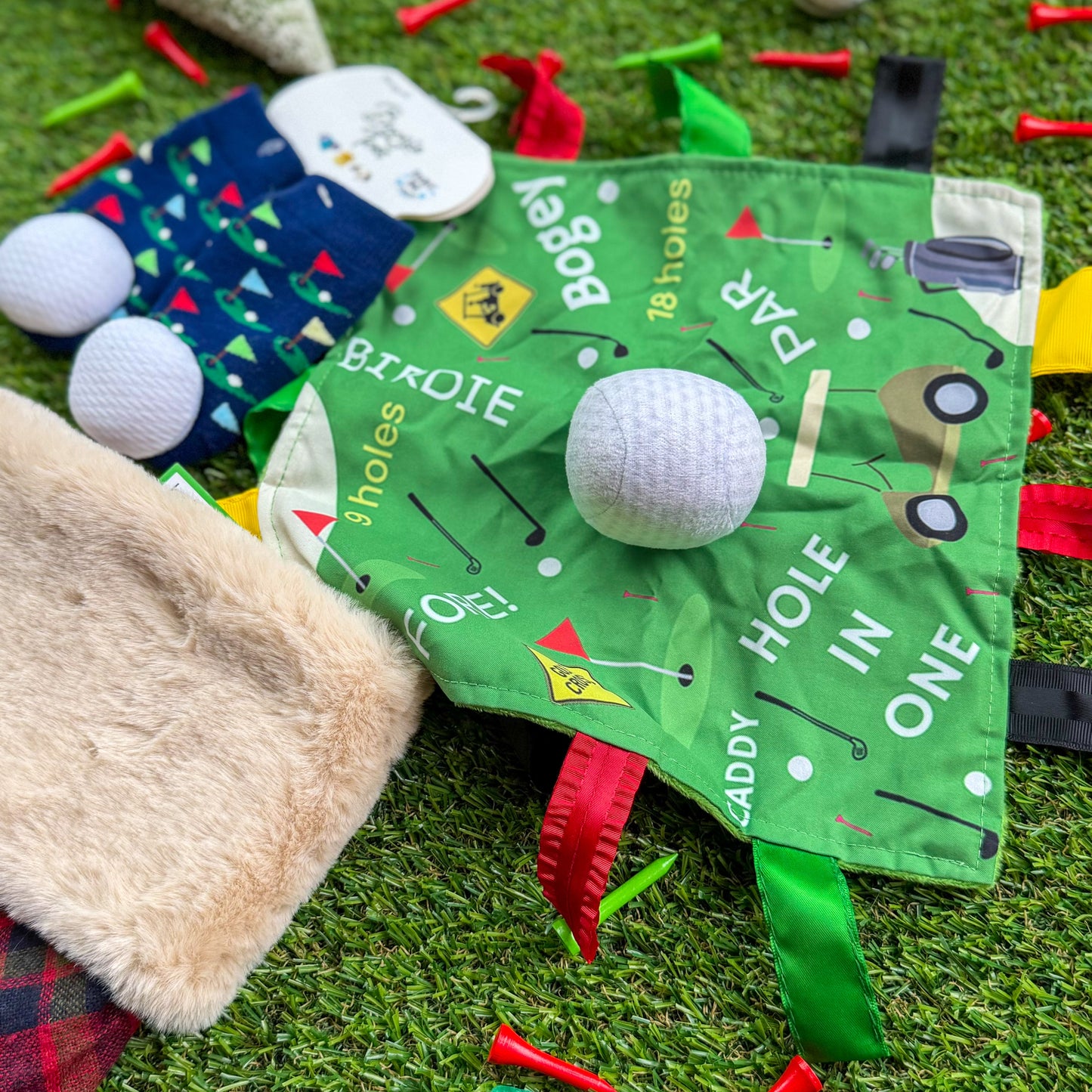 Golf-themed baby sensory lovey blanket with ribbon tags, paired with golf rattle socks and plush golf balls, displayed on artificial turf—ideal golf baby gift or stocking stuffer.