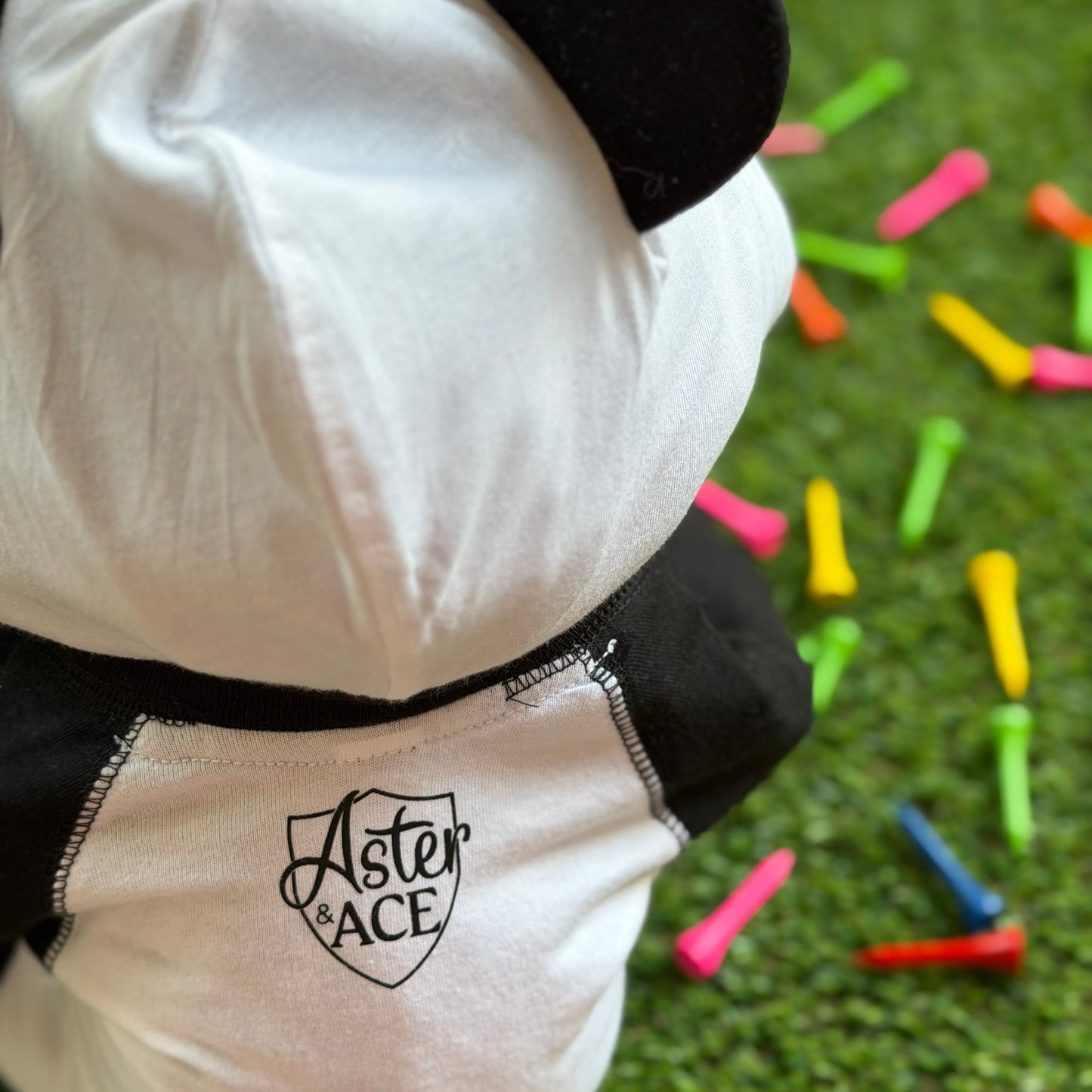 Close-up of the back of a baby hooded bodysuit with black bear ears and contrast stitching, featuring the Aster and Ace shield logo near the neckline, shown on grass with colorful golf tees.