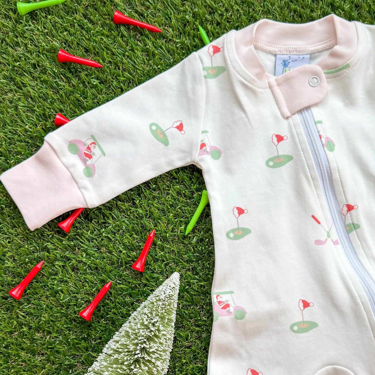 Pink-trim baby Christmas golf onesie by Aster and Ace featuring holiday golf cart and golf green print, shown on turf with red and green tees.
