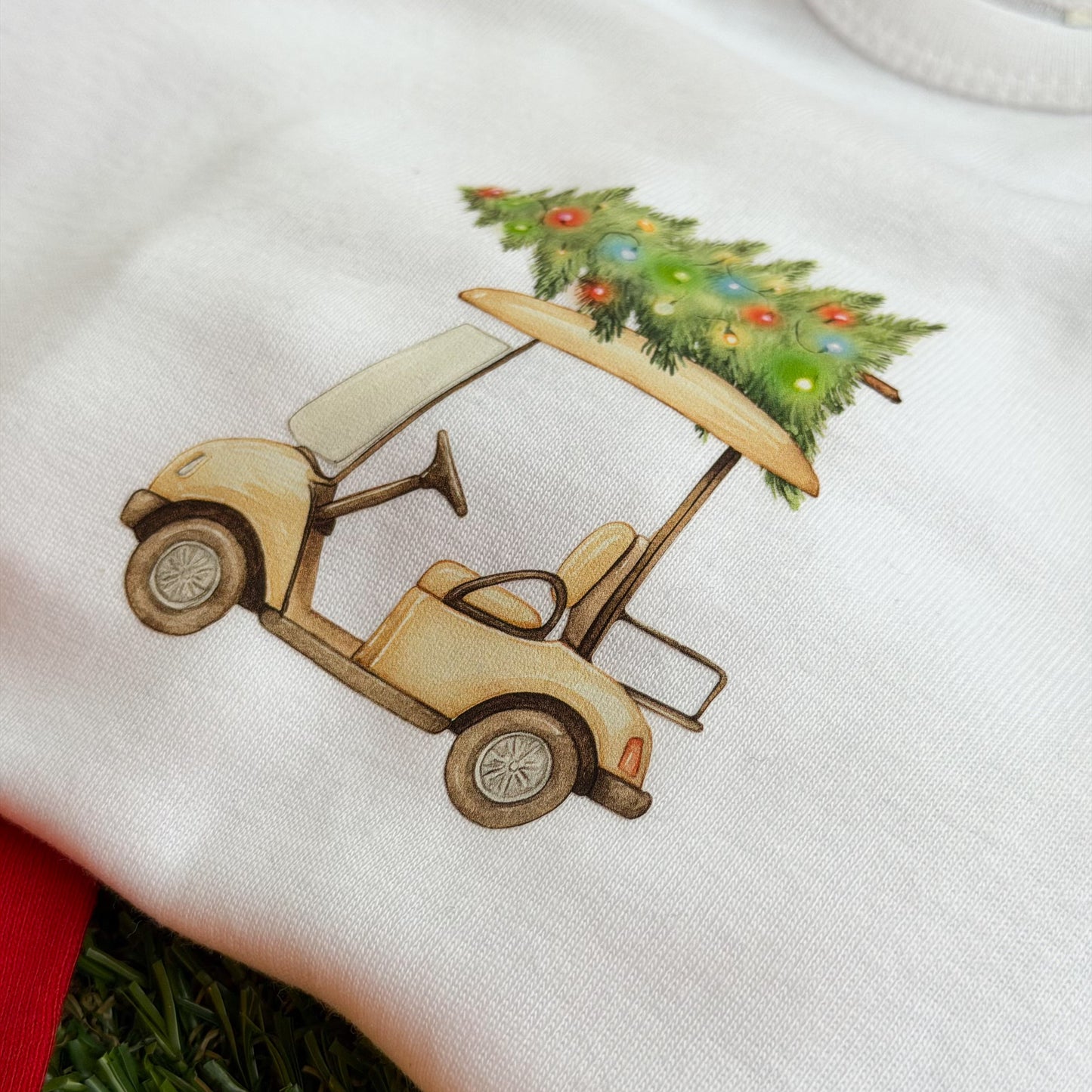White baby coverall with a festive golf cart graphic carrying a decorated Christmas tree, shown on green turf.