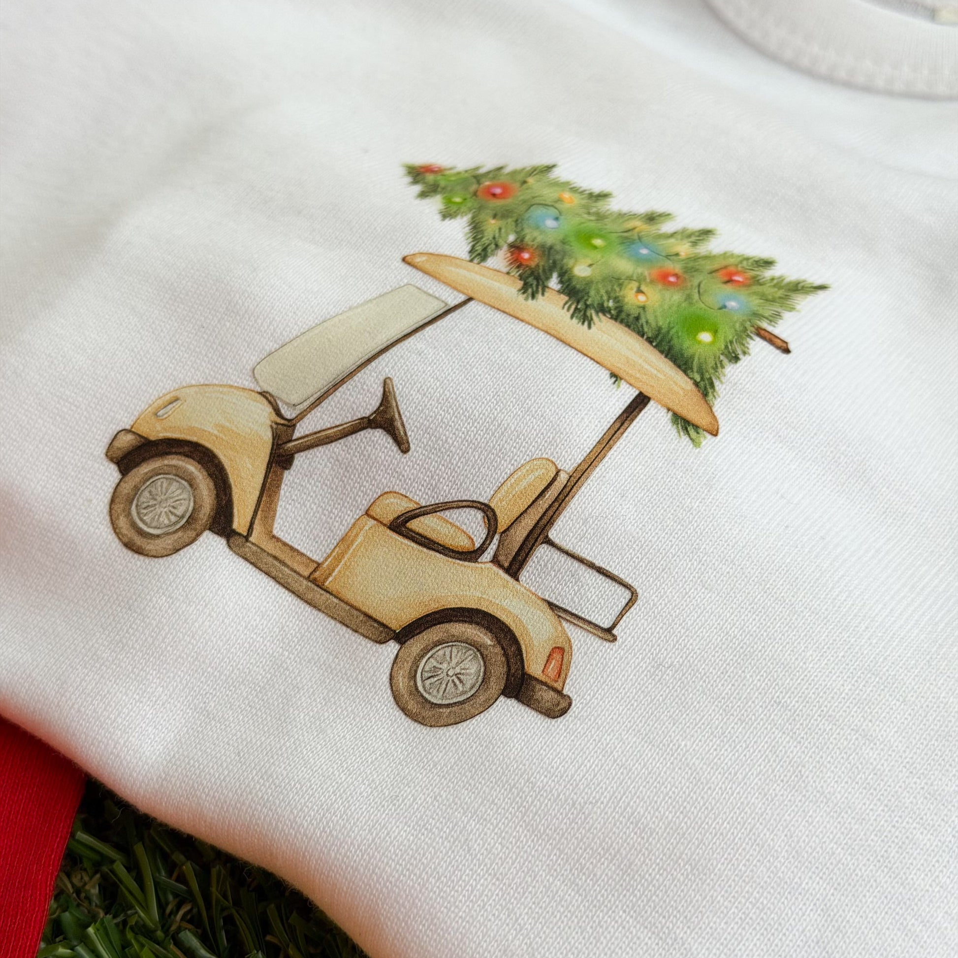 White baby coverall with a festive golf cart graphic carrying a decorated Christmas tree, shown on green turf.