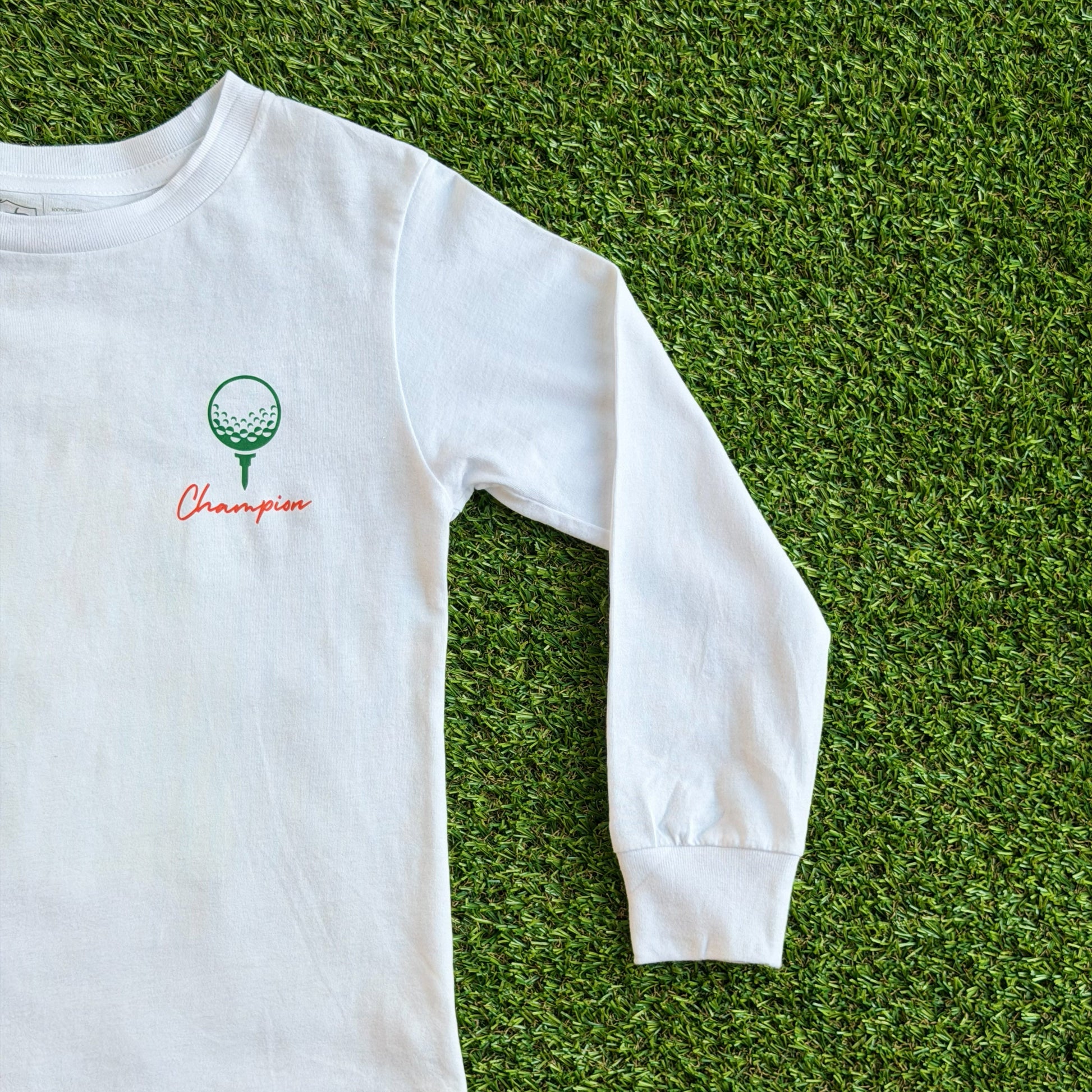Front of long sleeve white golf t-shirt featuring a small green golf ball on a tee graphic with the word “Champion” in red script beneath it, displayed on a grass background.