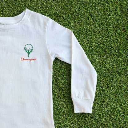 Front of long sleeve white golf t-shirt featuring a small green golf ball on a tee graphic with the word “Champion” in red script beneath it, displayed on a grass background.