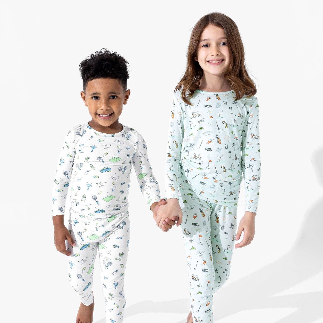 Two children wearing pajamas and holding hands. One has on white pajamas with a green and blue cartoon tennis print, the other has on light blue pajamas with multi-colored cartoon golf print.