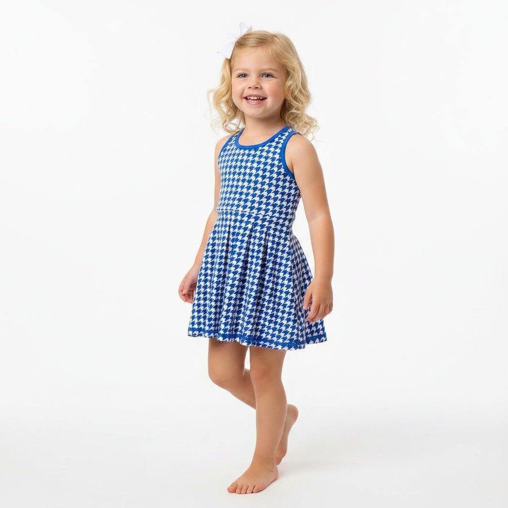 A young girl with blonde curly hair stands barefoot against a plain white studio background, wearing a sleeveless blue and white houndstooth active dress.