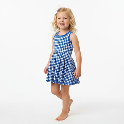 A young girl with blonde curly hair stands barefoot against a plain white studio background, wearing a sleeveless blue and white houndstooth active dress.