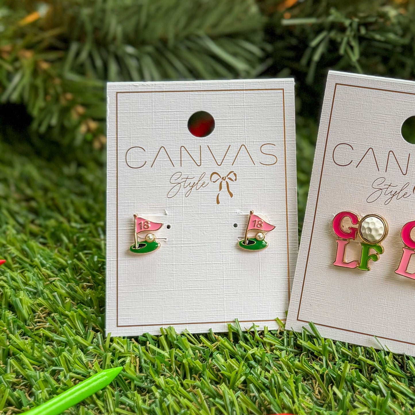 Canvas Style 18th-hole golf flag stud earrings displayed on product cards, shown on artificial grass with a holiday tree background—perfect golf-themed stocking stuffer jewelry for kids and adults.