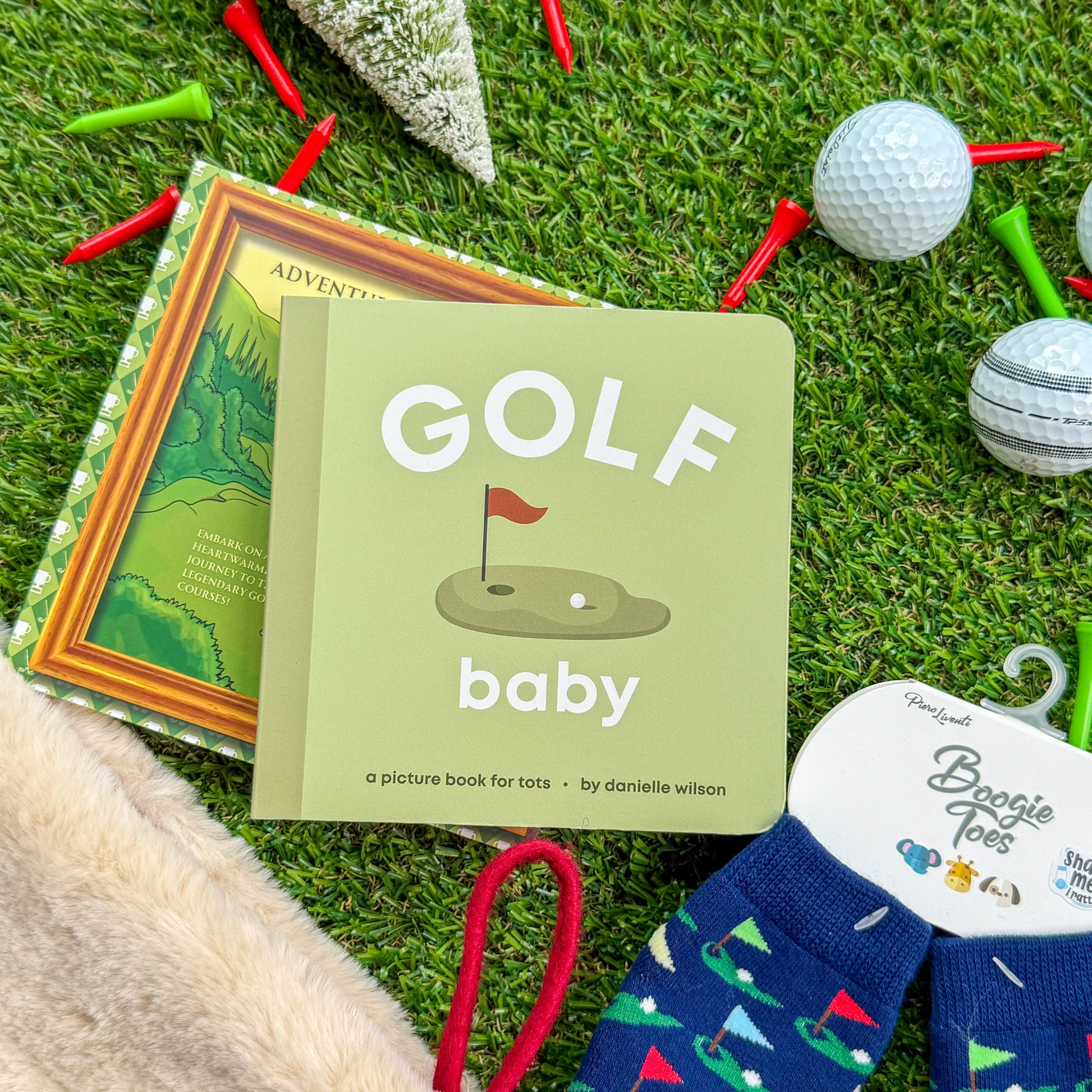 Golf-themed baby board book titled “Golf Baby” displayed with children’s golf rattle socks, holiday décor, and golf balls on artificial turf—perfect gift for little golfers and baby stocking stuffers.