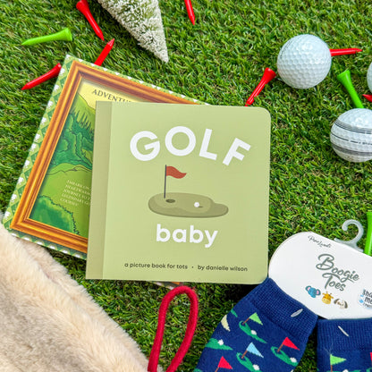 Golf-themed baby board book titled “Golf Baby” displayed with children’s golf rattle socks, holiday décor, and golf balls on artificial turf—perfect gift for little golfers and baby stocking stuffers.