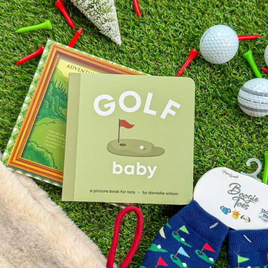 Golf-themed baby board book titled “Golf Baby” displayed with children’s golf rattle socks, holiday décor, and golf balls on artificial turf—perfect gift for little golfers and baby stocking stuffers.