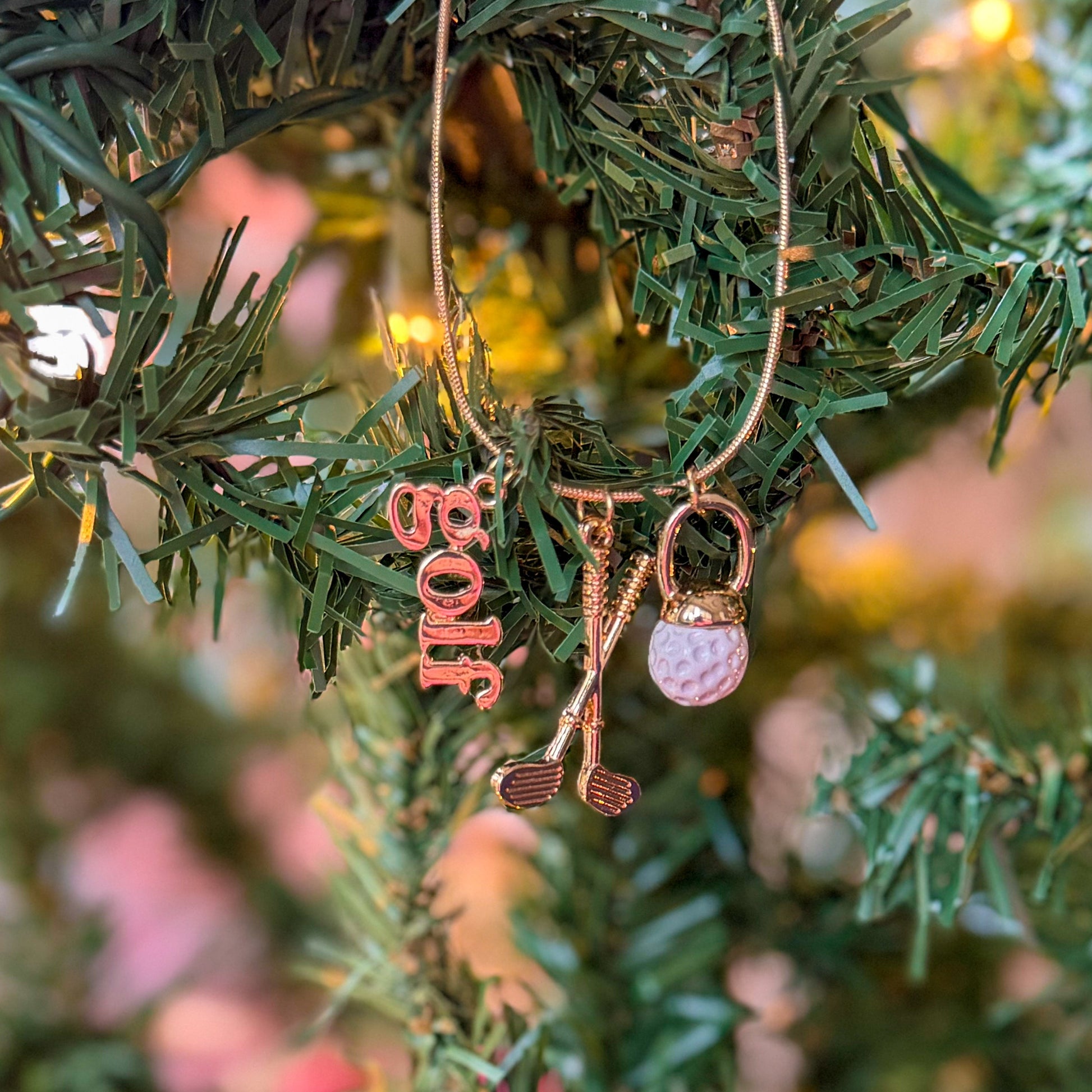Gold golf-themed necklace hanging on a Christmas tree branch, featuring charms shaped like a golf ball, crossed golf clubs, and a “golf” word pendant, styled with warm holiday lights for a festive gift look.