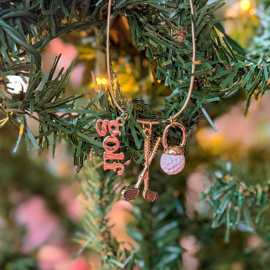 Gold golf-themed necklace hanging on a Christmas tree branch, featuring charms shaped like a golf ball, crossed golf clubs, and a “golf” word pendant, styled with warm holiday lights for a festive gift look.