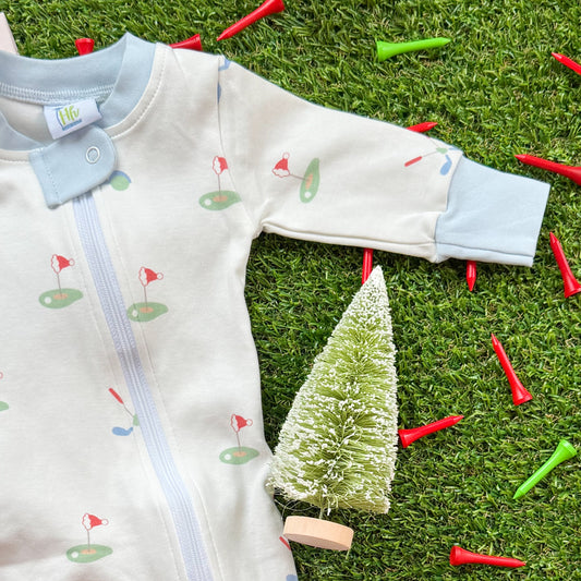 Close-up of blue-trim holiday golf baby onesie featuring Santa golf cart and golf green illustrations, shown on turf with festive tees.