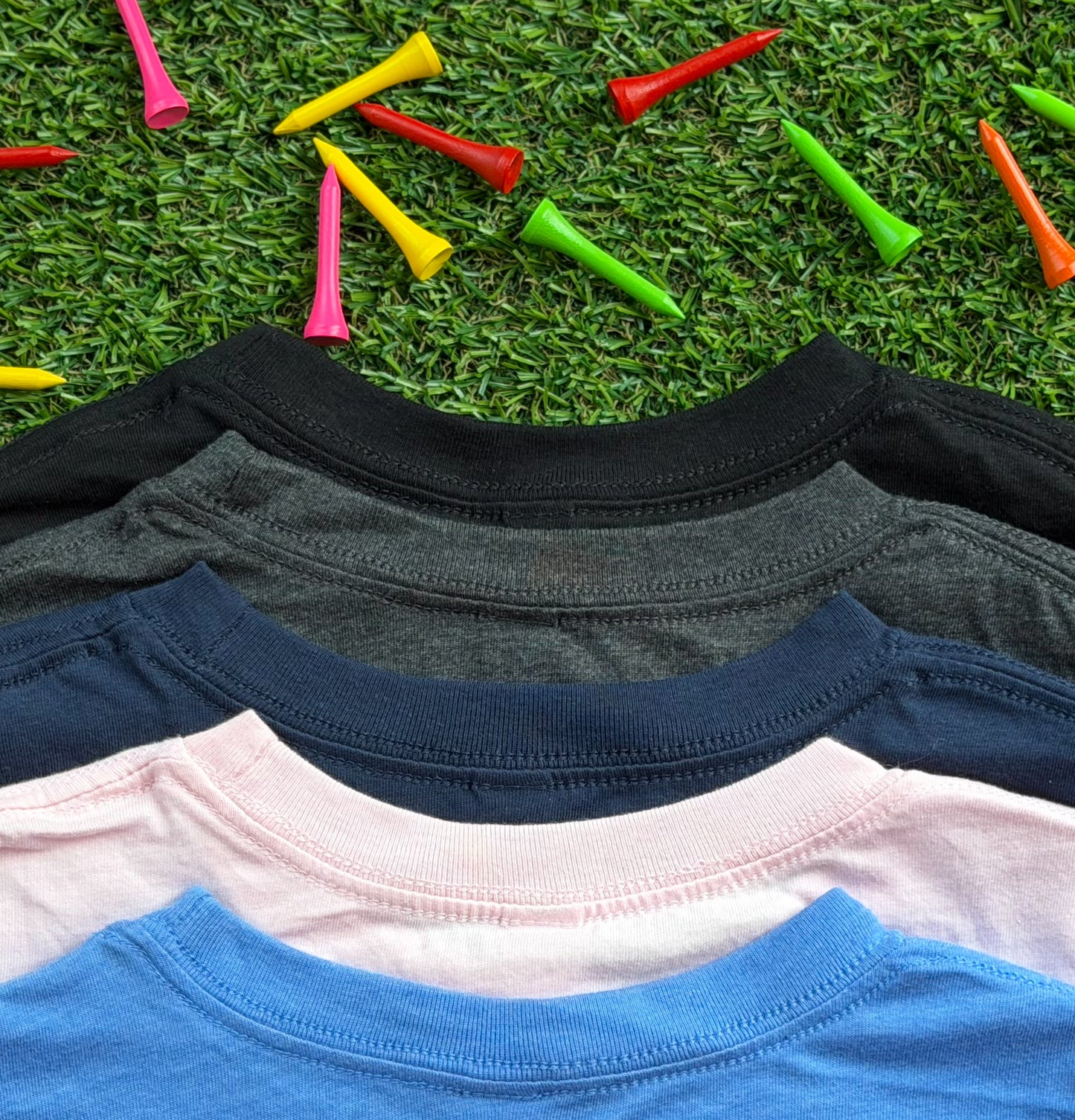 Close-up of the necklines of Royal-Tee long sleeve golf shirts in heather Columbia blue, pink, navy, heather charcoal, and black, displayed on green turf with colorful golf tees scattered around.