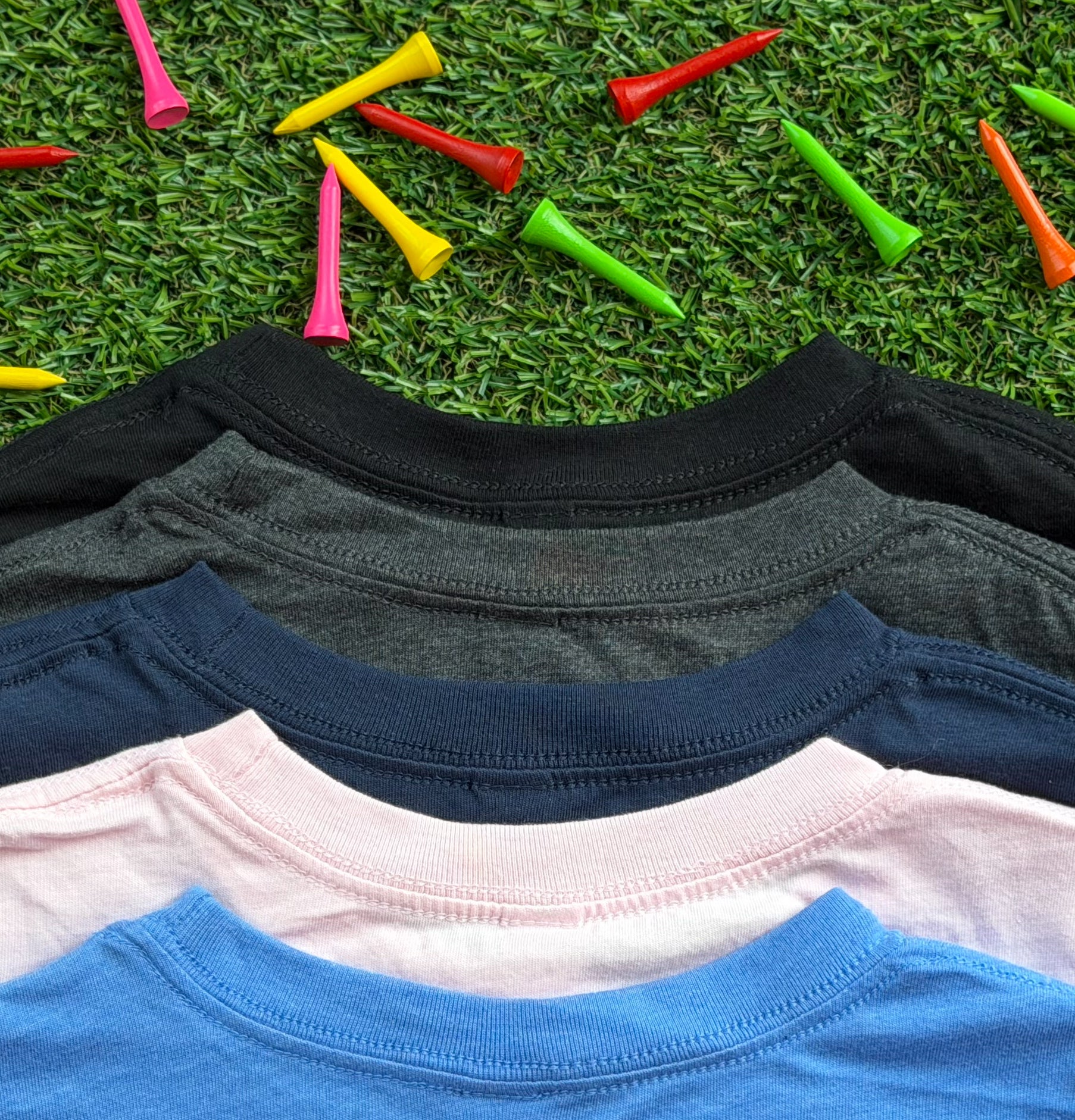 Close-up of the necklines of Royal-Tee long sleeve golf shirts in heather Columbia blue, pink, navy, heather charcoal, and black, displayed on green turf with colorful golf tees scattered around.
