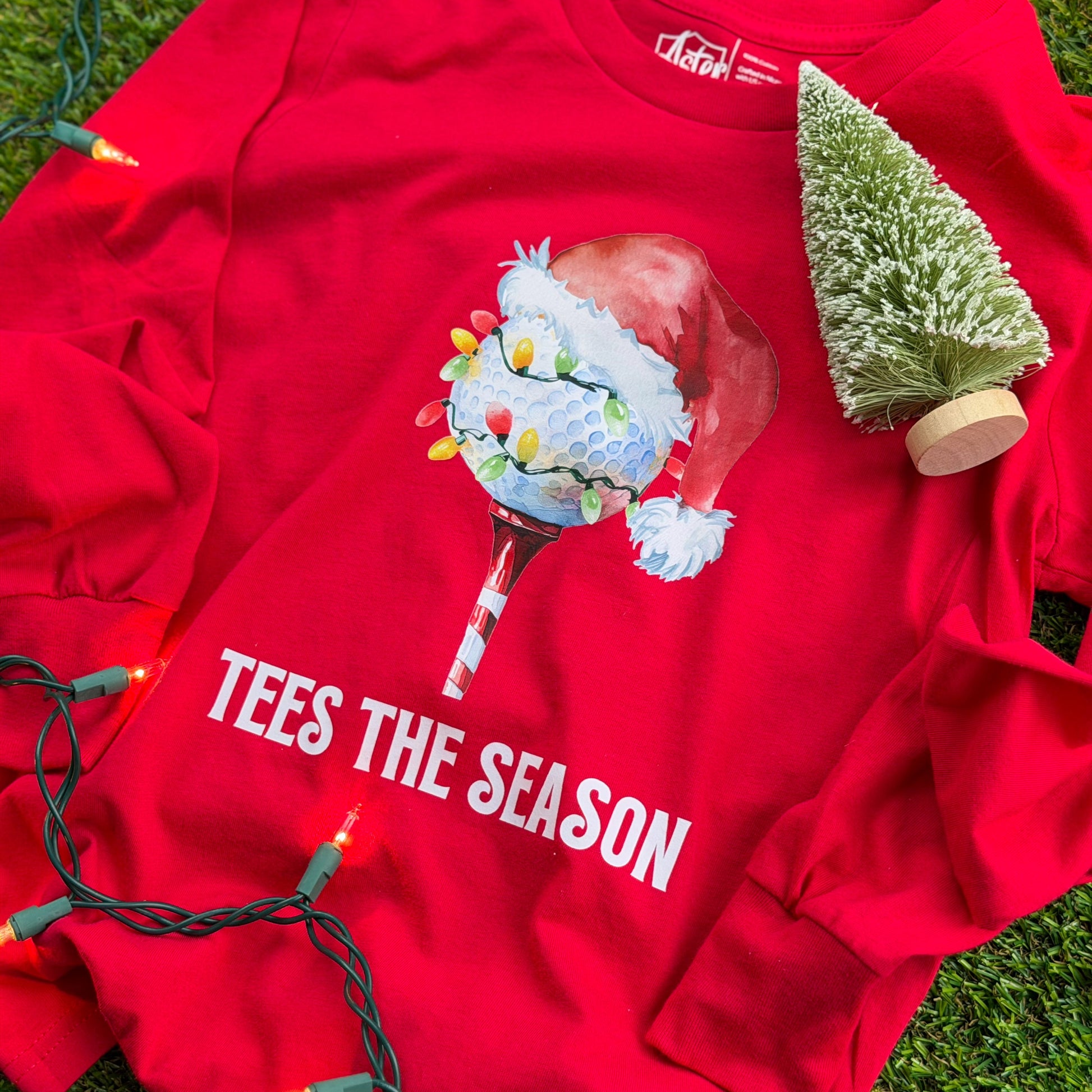 Red youth long-sleeve holiday golf shirt with a Santa-hat golf ball wrapped in Christmas lights and “Tees the Season” text, displayed on green turf with lights and a small tree.