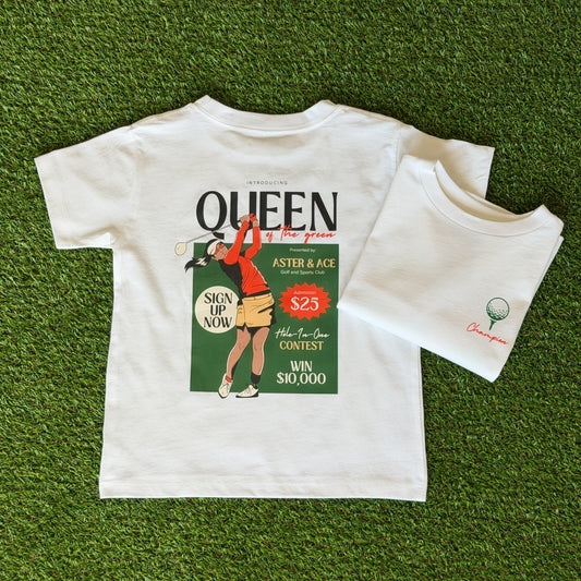 Queen of the Green white short sleeve t-shirt. The back of the shirt is pictured with large Queen of the Green graphic. Another white shirt is folded next to it to display the front design of the shirt. There is a green golf ball on a green tee with the word Champion written in orange script font.