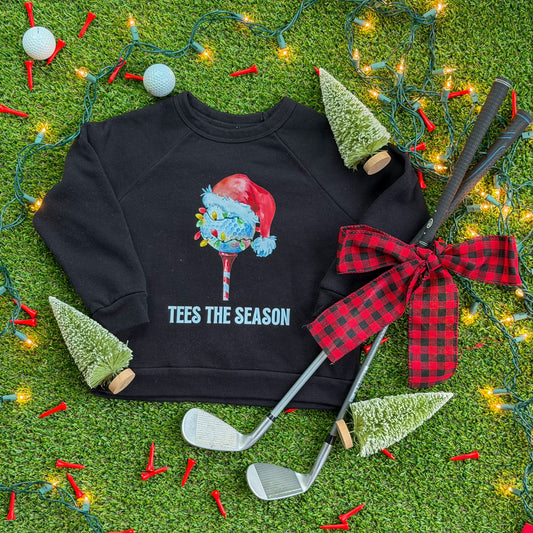 Black kids’ “Tees the Season” Christmas golf sweatshirt featuring a Santa hat golf ball graphic, displayed on turf with holiday lights, red golf tees, mini bottlebrush trees, and golf clubs tied with a red plaid bow.