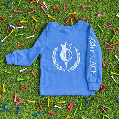 Front of kids’ long sleeve golf t-shirt in heather Columbia blue. Featuring the Aster + Ace “Royal-Tee” crest design with a crowned golf ball graphic on the chest and Aster and Ace wordmark logo down the left sleeve.