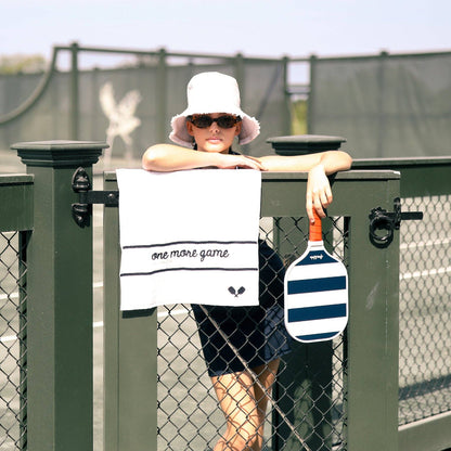 Child leaning on a tennis court fence with white embroidered towel hanging over the rail and a striped paddle beside it