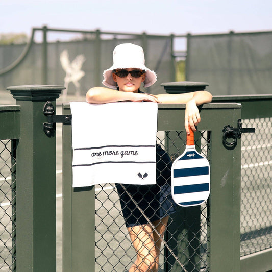 Child leaning on a tennis court fence with white embroidered towel hanging over the rail and a striped paddle beside it