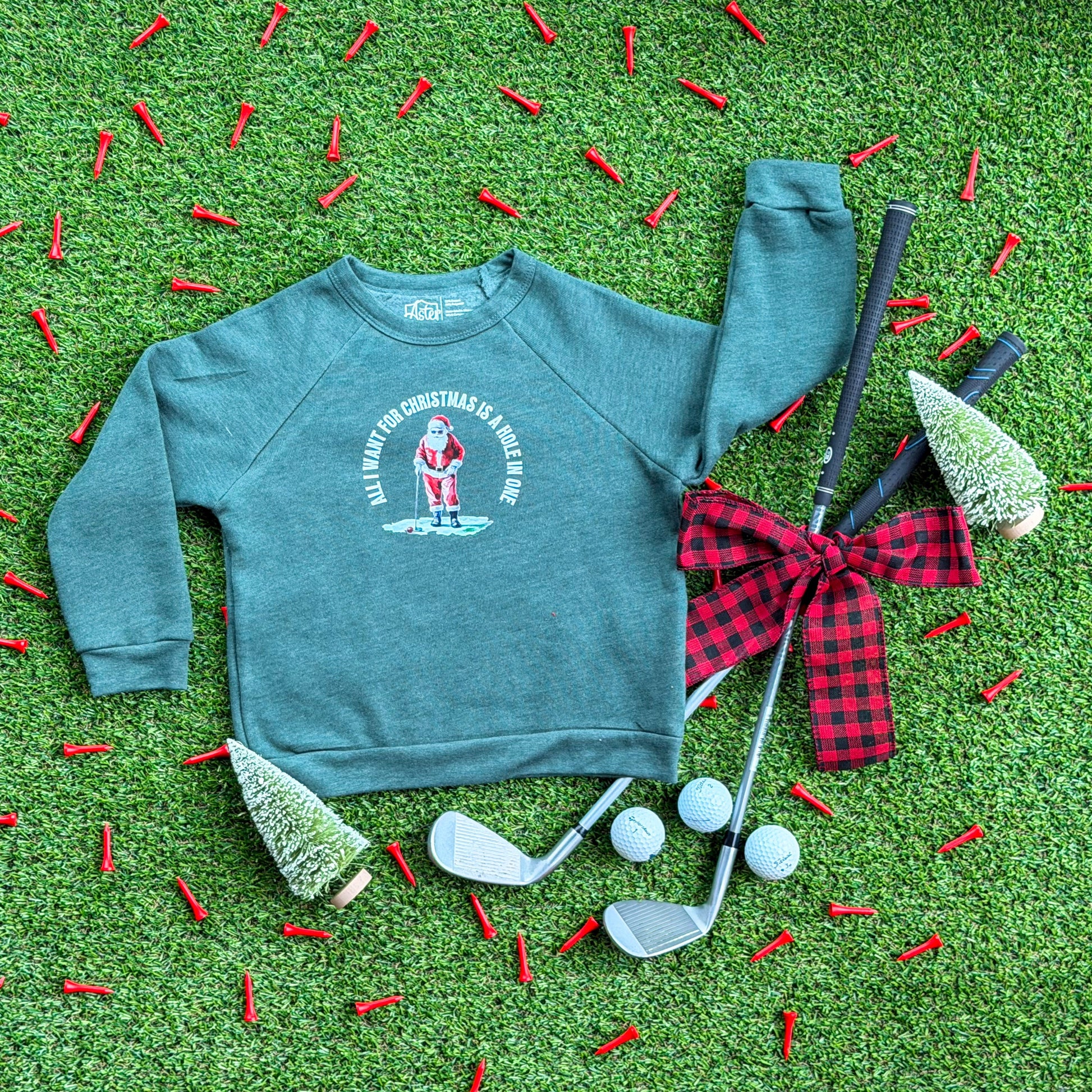Heather forest green kids’ Christmas sweatshirt featuring a Santa golfing graphic with “All I Want for Christmas Is a Hole in One,” styled on turf with golf tees, golf clubs, golf balls, and holiday décor.