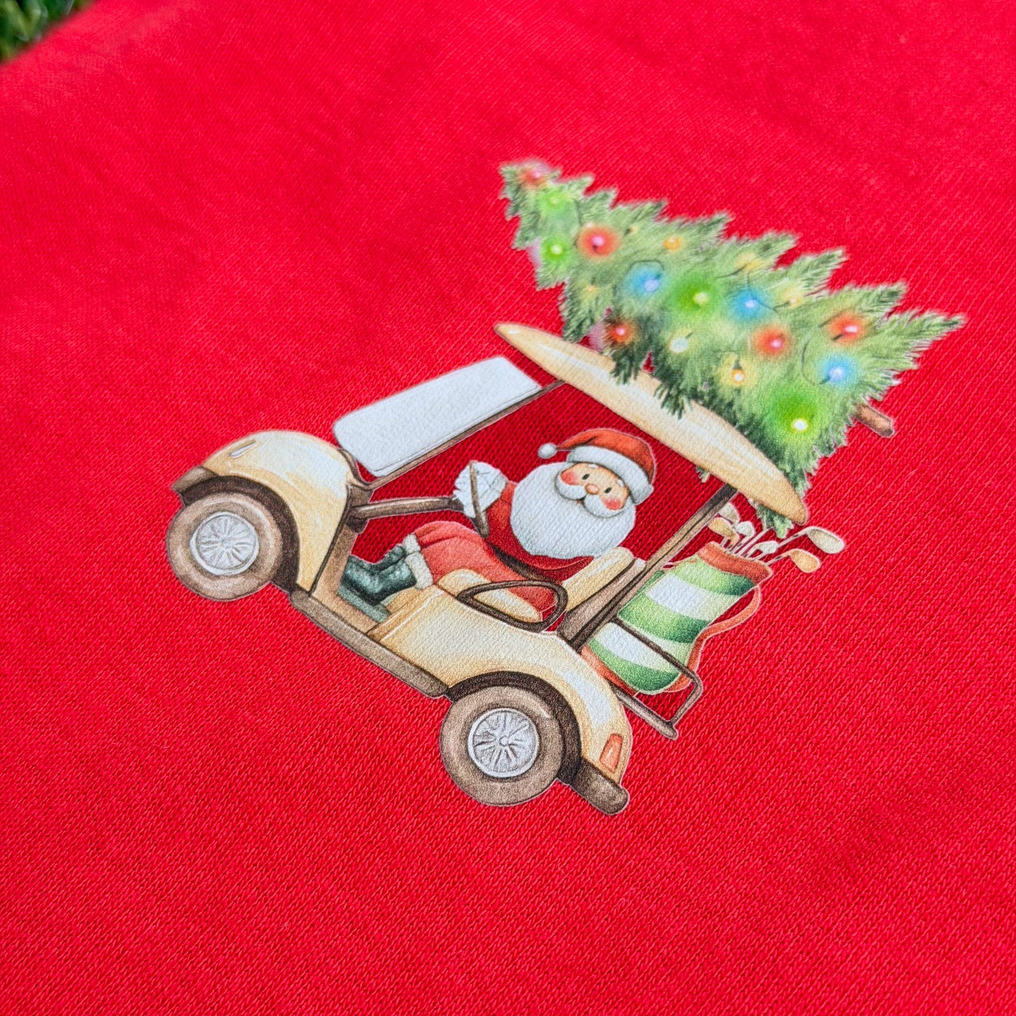 Red baby coverall featuring Santa driving a golf cart with a Christmas tree and holiday lights on top, shown in a close-up on fabric.