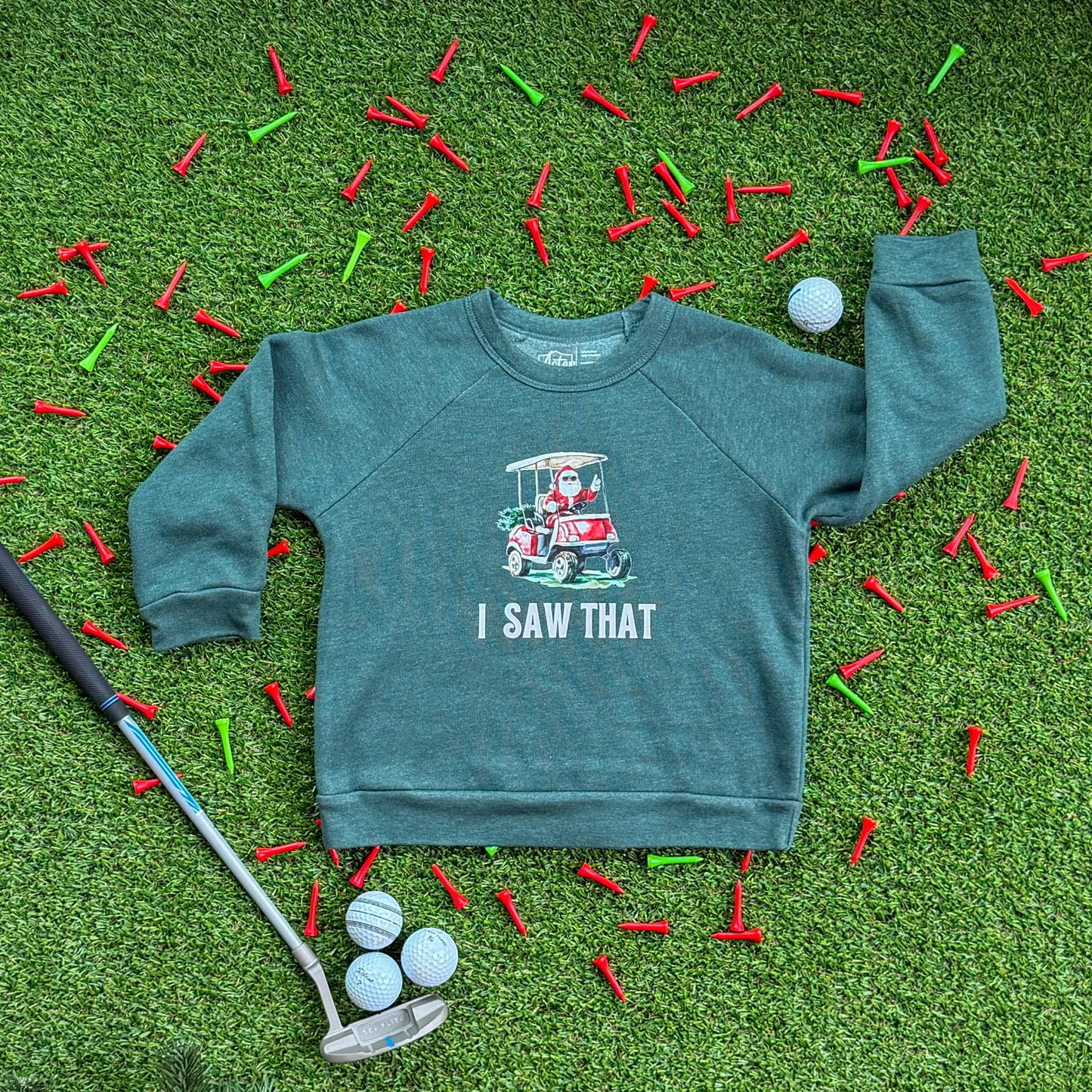Heather green kids’ sweatshirt with a Santa golf cart graphic and the text “I Saw That,” displayed on turf with red and green golf tees, golf balls, and a putter.
