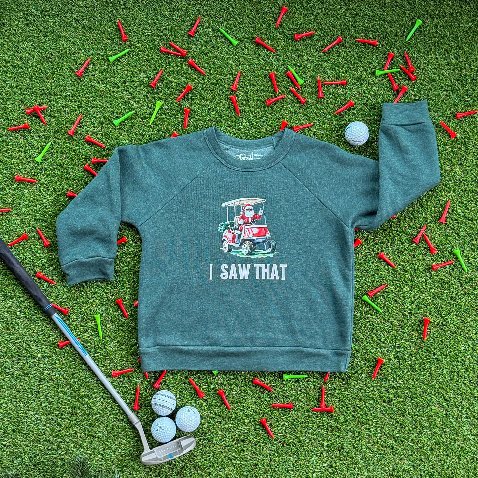 Heather green kids’ sweatshirt with a Santa golf cart graphic and the text “I Saw That,” displayed on turf with red and green golf tees, golf balls, and a putter.